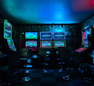 Dimly lit gaming arcade featuring a variety of brightly illuminated slot machines and video gaming screens. Neon signs and colorful displays create a vibrant atmosphere. The arrangement of gaming machines suggests an inviting space for entertainment, with chairs positioned for players.