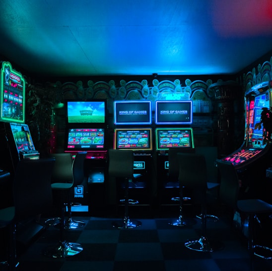 Dimly lit gaming arcade featuring a variety of brightly illuminated slot machines and video gaming screens. Neon signs and colorful displays create a vibrant atmosphere. The arrangement of gaming machines suggests an inviting space for entertainment, with chairs positioned for players.
