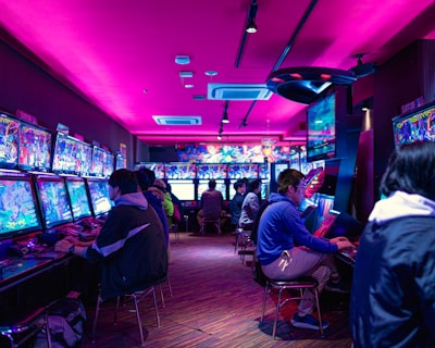 A vibrant arcade with rows of gaming machines adorned with colorful screens. Several people are immersed in playing games, seated on stools in a dimly lit room accented by an intense pink ceiling light. The atmosphere is lively and engaging.